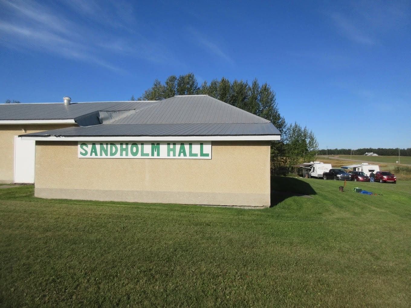 Tan building with green Sandholm Hall sign and grey metal roof on a grassy field.